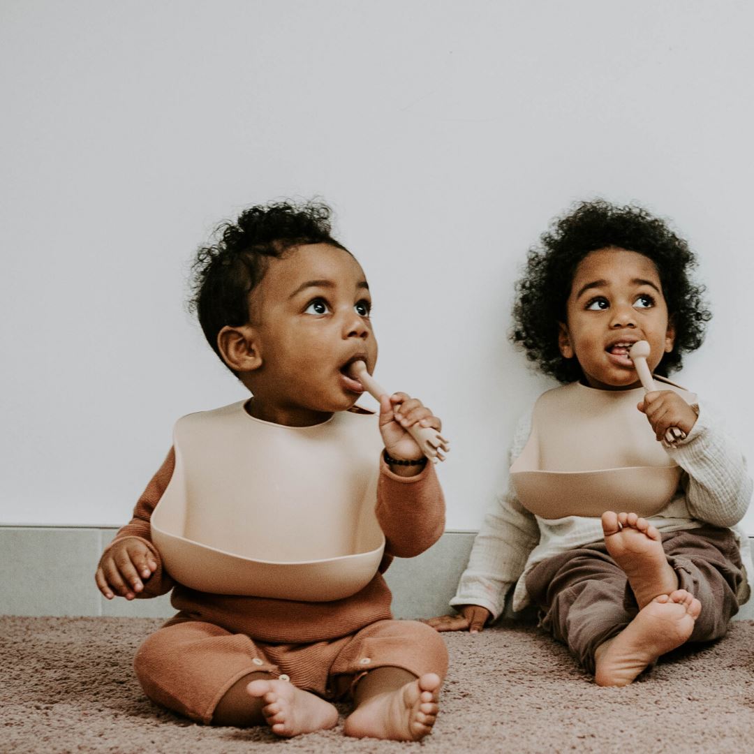 Toddler and baby wearing Boo Chew weaning bibs in Oat, chewing on spoons while sitting down