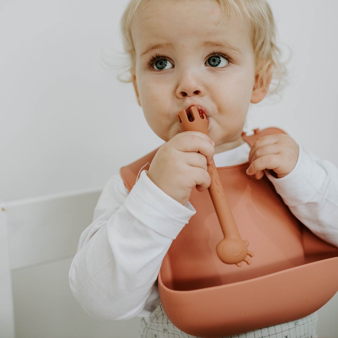 Silicone Feeding bib worn by boy aged 2 in a rust colour, feeding himself with Boo Chew 2 in 1 fork and spoon in rust colour