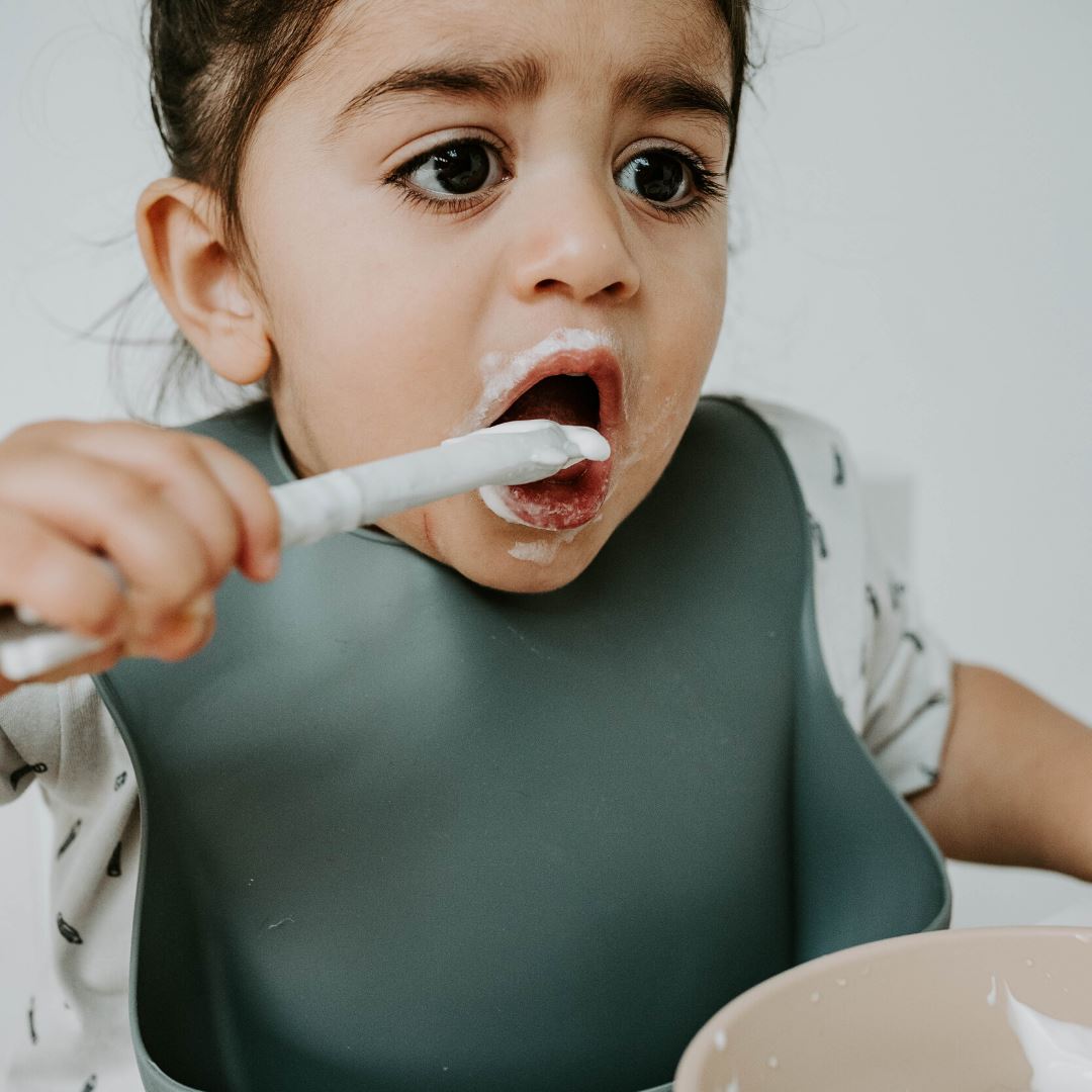 Toddler using baby's first utensil, feeding himself yoghurt with spoon head, marble coloured spoon, grey bib